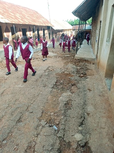 A group of children in matching maroon and white school uniforms walk along a cracked and uneven dirt path between two weathered, single-story buildings. The buildings have faded paint, corrugated metal roofs, and simple columns supporting the roof overhangs. The children appear to be heading in the same direction, possibly to or from school. The path is worn and uneven, showing signs of age and use. The overall scene depicts a rural or impoverished school environment.