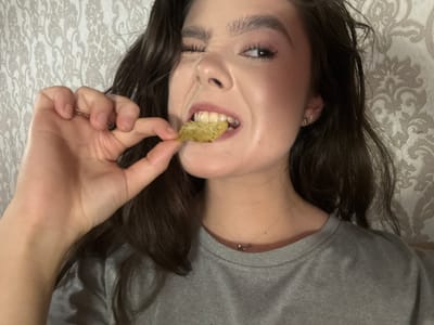The image is a close-up, chest-up shot of a young woman with dark, wavy hair and fair skin. She is wearing a gray t-shirt and a delicate necklace with two small rings. Her left eye is closed in a wink, while her right eye is open, looking towards the camera. She is holding a single, thin, rectangular-shaped chip, possibly a cracker or a vegetable chip, between the thumb and index finger of her left hand, bringing it to her mouth. Her teeth are slightly parted, and her lips are glossed. The background is a textured wallpaper with a light, ornate floral pattern. The lighting appears to be artificial and somewhat dim, casting shadows that highlight the contours of her face and hand. The overall impression is casual and playful. There is no discernible indication of the city of Zhovte, Ukraine within the image itself.