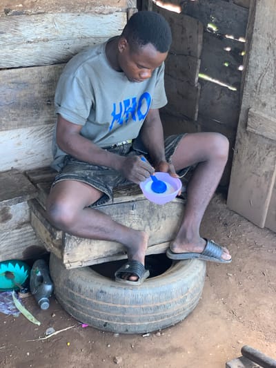 In Gbeleko, Nigeria, a young Black man is seated on a makeshift wooden structure composed of stacked planks, which is itself perched atop a worn vehicle tire. He is wearing a grey t-shirt with blue lettering and dark denim shorts. He is holding a light purple bowl with a blue spoon, appearing to be eating or preparing food. His feet are bare except for black slide sandals. The surrounding environment consists of rough, wooden walls, suggesting a rustic or unfinished structure. Debris, including plastic bottles and what appears to be a green plastic item, is visible on the dirt ground around the tire. The scene is illuminated by natural light filtering into the structure.