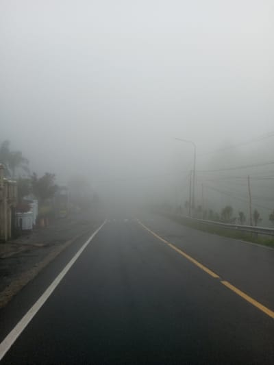A fog-laden road stretches into the distance, its asphalt glistening wetly. A solid white line marks the left edge, while a dashed yellow line runs along the road's center. Barely visible buildings and trees line the left side, obscured by the dense fog. On the right, a metal guardrail flanks the road, with sporadic vegetation beyond. Power lines and a street lamp are dimly visible through the heavy fog. The overall atmosphere is eerie and visibility is severely limited.