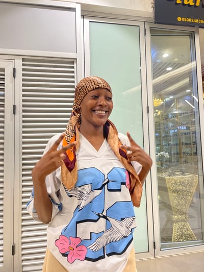 A young Black woman stands in front of a storefront in Unguwar Sarki, Nigeria. She is wearing a light-colored jersey with a blue, white, and pink graphic print featuring cranes and flowers. A patterned headscarf, in shades of brown and orange, is tied around her head, with a corner of the fabric draped over her shoulder. She is smiling broadly and making a peace sign with both hands. The storefront behind her has large glass windows and white paneled doors. Inside the store, shelves stocked with small bottles are visible, along with decorative gold-colored items. A sign with the phone number 0806248394 is also visible. The lighting suggests it is daytime.