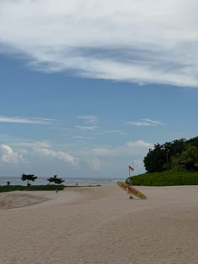The image depicts a beach scene in Nusa Dua, Indonesia. The sky is predominantly blue with wispy clouds. On the right side of the frame is a sandy beach with sparse vegetation. Adjacent to the vegetation, a yellow surfboard with the words "SURF RESCUE" is visible, along with a red and yellow flag. The ocean is visible as a narrow strip in the middle of the image. The scene is peaceful and suggests a tropical, coastal environment.