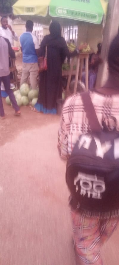 The image depicts a bustling scene at a market in Kabala, Nigeria. In the foreground, a person with a black backpack stands with their back to the camera. The backpack has white lettering and a logo. The person wears a plaid shirt and patterned pants.
Further into the scene, a woman in a long, black robe and head covering is at a fruit stand. The stand has bananas and other fruit. Nearby, several men stand by a pile of green watermelons. The setting is outdoors, with tents providing shade. The overall scene suggests a typical market day in Kabala.