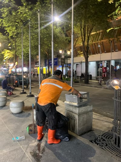 At night in Yogyakarta, Indonesia, a male sanitation worker is observed on a brightly lit sidewalk. He wears an orange long-sleeve shirt with "SODOBORO" and additional text, black cargo pants, bright orange rubber boots, a dark baseball cap, and a black face mask. His back is mostly to the viewer as he empties the contents of a grey square public trash receptacle, mounted on a stone base inscribed with "jogja," into a black trash bag positioned on the ground next to his left boot. A blue plastic water bottle lies horizontally on the sidewalk near his right foot, which is also close to a long-handled brush or broom.
The sidewalk features multiple light-colored, spherical concrete bollards and a decorative cast-iron grate for drainage. Directly behind the worker, a row of tall, thin silver poles extends into the background. Lush green trees, illuminated by streetlights, line the thoroughfare. In the background, other individuals are visible on the sidewalk and street, which appears busy with parked motorcycles, including a three-wheeled vehicle on the left, and brightly lit storefronts with various signs, one partially legible as "TONGCHAT." Buildings with distinctive brown-patterned facades are visible further down the street.