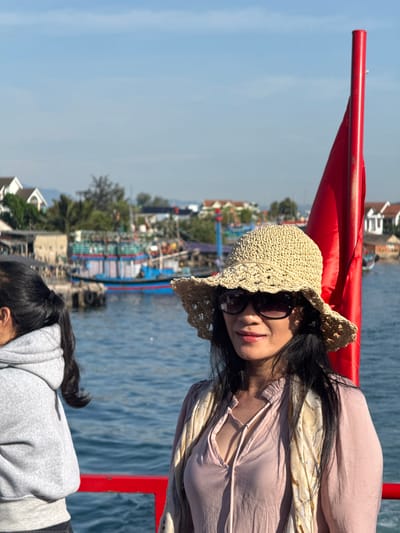 In An Hai, Vietnam, a woman wearing a straw hat and sunglasses is in the foreground, looking to her right. She is wearing a light pink blouse and a patterned scarf draped around her shoulders. Behind her, another person in a grey hooded sweatshirt is visible from the side. A tall, bright red pole stands prominently on the right side of the frame, partially obscuring a red railing. The background features a body of water, likely a harbor, with several colorful fishing boats docked. Buildings and trees line the distant shore under a clear blue sky.