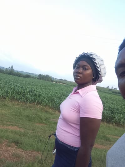 This media file captures a young woman with dark skin, wearing a pink polo shirt and a patterned head covering, standing in a grassy field. She is positioned on the right side of the frame, with her body angled away from the camera. Her gaze is directed towards the camera, and she has a neutral expression. A portion of another person's head, cropped from the right edge of the image, is also visible.

Behind the woman, a vast field of tall green crops, likely corn, stretches into the distance. The field is bordered by a line of trees and further hills in the background, under a pale, overcast sky. The overall impression is of an outdoor setting, possibly a rural area in Nigeria, given the location context. The lighting suggests it is daytime, though the cloudy conditions create a diffused light. The scene appears calm and uneventful. There is no visible text in the image.