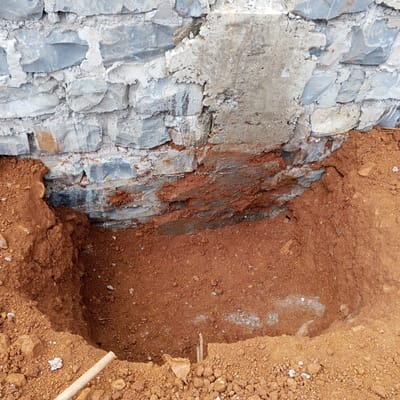 The image depicts a hole dug in the ground next to a stone foundation wall. The soil is reddish-brown. The stone wall is made of gray and white stones and mortar. There is a patch of exposed concrete on the wall. A wooden handle is visible in the lower left corner of the hole. Two sticks are in the hole. The image may have been taken in the Borabu Chitago ward of Kenya.