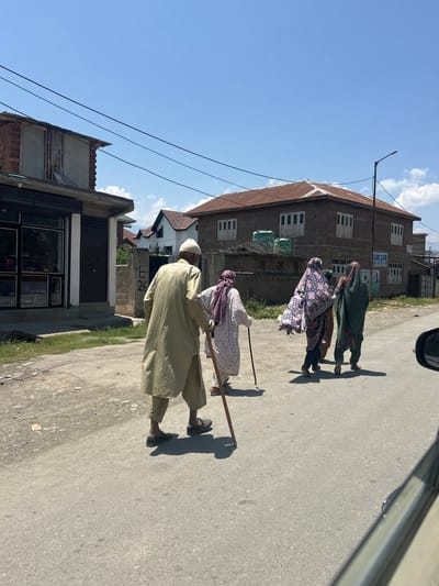 In Vessu, India, on a sunny day with a clear blue sky, four individuals are walking on a paved road. An older man, wearing a light-colored tunic and pants and a white cap, walks from left to right, leaning on a wooden cane. Behind him, three women, two of whom are veiled, are also walking in the same direction. The woman in the middle wears a patterned shawl over her clothes, and the woman to her right wears a dark green garment. The environment features buildings with corrugated metal and tiled roofs, as well as power lines. The foreground shows the edge of a vehicle.