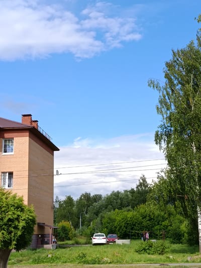 This is a daytime, outdoor, medium-shot photograph taken in Verea, Russia. The scene is set on a sunny day with a clear blue sky and scattered white clouds, suggesting fair weather. The foreground features a grassy area with a dense green evergreen tree on the left. Behind this, a multi-story brick building with a brown metal roof is visible, featuring several windows. A small covered entryway with a pink support structure is also present.
In the middle ground, a row of trees and lush green foliage forms a backdrop. A clothesline with several items of clothing hanging to dry is set up in front of the trees. Two cars, a white taxi and a red sedan, are parked in the grassy area to the left of the clothesline. In the background, more trees and a street light can be seen.
A tall birch tree with green leaves stands on the right side of the frame, partially obscuring the sky. Electrical wires are strung across the middle of the image, supported by poles. The overall atmosphere of the image is peaceful and depicts a typical residential scene. No text is visible in the image.