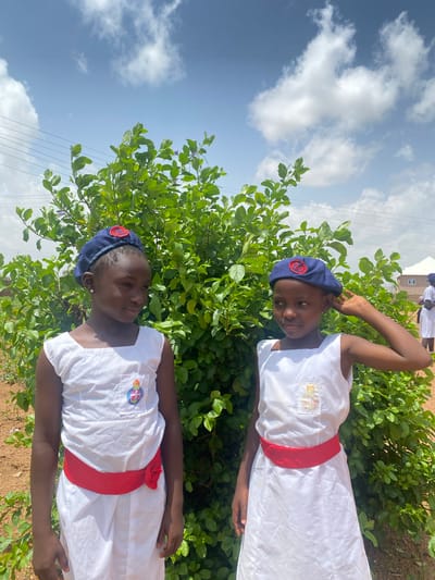 Two girls stand side-by-side in front of a leafy green shrub, against a backdrop of a light blue sky with scattered clouds. Both girls wear matching white dresses with a red sash around the waist and blue caps. A building can be partially seen in the background behind the shrub. The location is Turu, Nigeria.