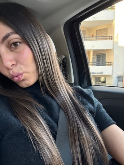 The image displays a female individual, observed from the chest up, seated in the front passenger position of a vehicle. She has long, straight, dark brown hair with lighter brown highlights, brown eyes, and fair skin, with her lips intentionally puckered. She wears a dark, possibly black or charcoal, turtleneck sweater and is secured by a black seatbelt across her chest.

Through the vehicle's window, an exterior environment is visible under daylight conditions. A multi-story building, light-colored (beige/cream), with multiple dark-railed balconies, is prominently featured. A spherical, reddish-orange object rests on an upper balcony. On the building's facade, a white sign with black lettering displays "taux.com".

This building is specifically located on Avenue Pasteur in Antibes, France, where "Taux.com" is associated with real estate and financial services. The individual is looking towards the camera, suggesting a posed photograph.