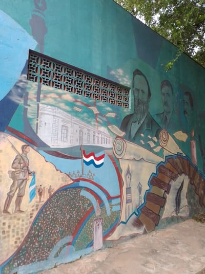 The image shows a mural on a wall in Villa Hayes, Paraguay. The mural features several elements: portraits of three men (likely historical figures), a depiction of a building (potentially a significant landmark in Villa Hayes), a soldier carrying a flag, a flag of Paraguay, stylized quills and scrolls, and lit candles. The mural is painted in various shades of blue, green, brown, and white. Above the mural is a decorative lattice-like window vent. The background shows trees. The mural is on the side of a concrete structure with pavement in the foreground.
