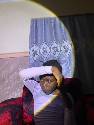 A young Black female with dark hair pulled back sits on a red couch in an indoor setting, possibly a home in Barakin Sabawa, Nigeria. A dramatic beam of yellow and white light casts a sharp shadow across her face, torso, and the surrounding area, illuminating the patterned blue curtain behind her and the splattered paint on the wall to her left. She is wearing a long-sleeved, light purple shirt with a white panel on the chest. Her right arm is bent, with her hand resting on her forehead, as if shielding her eyes or in contemplation. The couch upholstery features a dark red background with black floral patterns. The lighting creates a stark contrast between illuminated and shadowed areas of the scene.