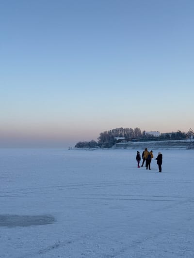 The image depicts an outdoor winter scene on a frozen body of water in Liutizh, Ukraine, under a clear, gradient sky transitioning from light blue overhead to a pale pink-orange near the horizon. The foreground and midground consist of a flat, extensive expanse of snow-covered ice, marked by linear tracks. In the lower-left foreground, an irregular, dark-toned patch, consistent with open water or thin ice, is visible. Four individuals are present in the midground, standing on the frozen surface in winter clothing. One individual wears a bright yellow jacket and dark pants, with one leg lifted, suggesting movement or preparation for ice skating. The background reveals a snow-covered shoreline featuring multiple bare trees, several buildings with visible roofs, and a low retaining wall. A short, dark-colored pier-like structure extends from the shoreline into the frozen water. Natural daylight illuminates the scene uniformly.