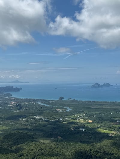 This is a high-angle aerial view of a coastal area in Ban Din Daeng Nui, Thailand. In the foreground, dense green forests cover rolling hills, with sunlight casting shadows on the foliage. Further in the midground, a mix of lush green agricultural fields, possibly plantations, and scattered residential buildings are visible. A winding river or canal cuts through this landscape, leading towards the coast. The shoreline is lined with trees, and beyond that lies a large body of turquoise water. Several limestone karst islands, characteristic of the region, emerge from the sea. In the distance, a hazy horizon suggests more islands and landmasses. The sky is a bright blue with scattered white clouds, some of which are casting shadows on the land below. The overall scene depicts a tropical coastal environment with a blend of natural landscape and human settlement.