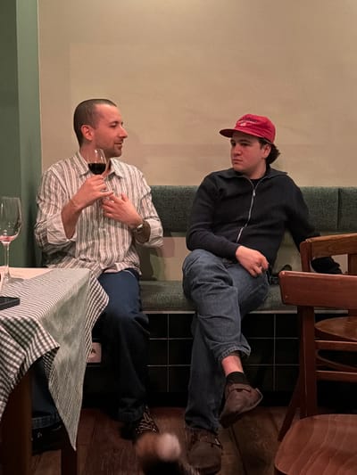 The image depicts two adult males seated on a dark green upholstered booth in an indoor dining establishment in Ixelles - Elsene, Belgium. The background consists of light beige walls above the booth and dark tiled panels below. Wooden flooring is visible at the bottom.

On the left, a table with a green and white checkered tablecloth is partially visible, holding an empty stemmed wine glass and a dark mobile phone.

The male on the left has short, dark hair and wears a vertical striped long-sleeved button-up shirt in light and dark tones, dark blue trousers, and dark-colored shoes with lighter laces. A watch is visible on his left wrist and a bracelet on his right. He holds a stemmed wine glass containing red liquid in his left hand and gestures with his right hand towards his chest. He is turned towards the male on his right, appearing to speak, with a slight smile.

The male on the right wears a red baseball cap with a white emblem, a dark blue/black zip-up jacket, blue jeans, dark socks, and brown slip-on shoes. He is seated with his legs crossed, with his hands resting on his lap. He is looking towards the male on his left with a neutral expression. A wooden chair with a brown seat is partially visible on the far right. A blurry, light-colored object is visible near the bottom center of the frame.