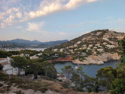 This is a landscape photograph taken from an elevated vantage point overlooking a bay in the city of Andratx, Spain. The scene is dominated by a hillside covered in dense green trees and dotted with numerous white buildings, indicative of a residential area. The buildings are modern in design, with flat roofs and large windows, and appear to be constructed on terraces carved into the hillside.
In the foreground, the terrain is covered with dry, sparse vegetation, suggesting a Mediterranean climate. The mid-ground features a secluded cove with clear, blue-green water. A white sailing yacht is anchored in the cove. Rocky outcrops and more trees line the edge of the water.
In the background, a range of hazy, blue mountains forms the horizon. The sky is partly cloudy, with soft white clouds scattered across a pale blue expanse. The overall impression is one of a peaceful coastal settlement nestled amongst natural beauty. There are no visible people or animals in the frame.