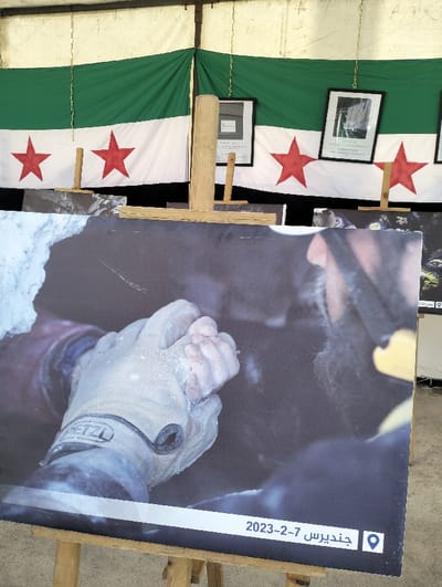 This image documents an exhibition display located within the city of Homs, Syria, set within a structure featuring draped beige and white canvas material. The background presents two horizontally oriented flags, each displaying a green stripe, a white stripe with three red stars, and another green stripe, consistent with the flag of the Syrian opposition. Several framed photographs hang by chains against these flags, partially obscured by various wooden easels. In the foreground, a prominent large-format photograph is positioned vertically on a wooden easel. This photograph depicts a close-up view of a gloved hand, marked with a 'PETZL' logo, firmly grasping an ungloved hand covered in light-colored debris. The context within the photograph suggests a rescue scenario in a dark, confined space, likely a collapsed structure. A white banner along the bottom edge of this large photograph contains the date '2023-2-7' and the Arabic word 'جنديرس' (Jindires) accompanied by a location pin icon, indicating the geographic origin of the depicted event.