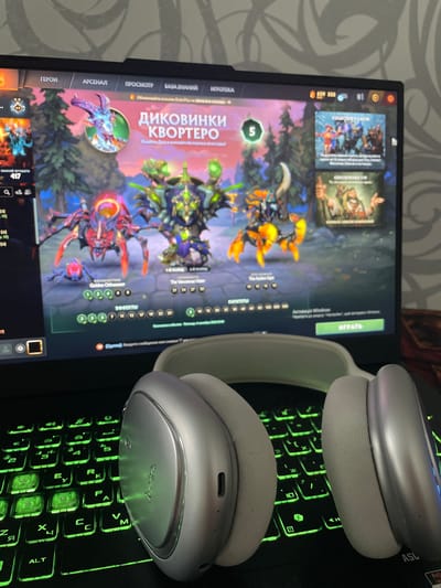 The image shows a close-up of a laptop screen displaying the game Dota 2. The game interface is visible, with character models and text in Russian. To the right of the screen, there is a section labeled "COLLECTOR'S CACHE" and "ОБНОВЛЕНИЕ 7.36". Below the laptop, a pair of silver-colored headphones with light gray earcups rests on a keyboard that is illuminated with green backlighting. The overall scene suggests a person is about to play or is currently playing a video game. The location of Rozsoshentsi, Ukraine is not depicted or suggested in the image.