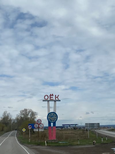 The image captures a roadside scene near Oyok, Russia, marked by a prominent entry sign displaying "OËK" in red Cyrillic characters above a blue circular emblem with Cyrillic text. The sign is flanked by horizontal red and blue stripes, and positioned near a collection of traffic signs regulating weight and vehicle types, as well as a diamond-shaped warning sign. The foreground shows a paved road with a white dividing line, bordered by patches of green vegetation and bare trees. The background features a cloudy sky. A road with several signs runs in the distance.