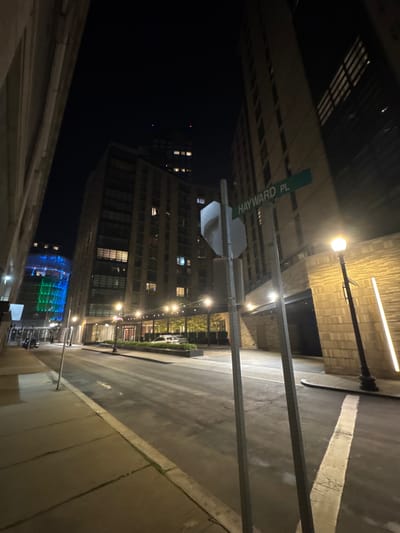 The image displays a nighttime urban street scene on Hayward Place in Boston, Massachusetts, United States. The perspective is low-angle, looking down the street framed by tall, multi-story buildings.
In the immediate foreground, two metallic posts support a green street sign with "HAYWARD PL" in white text, angled towards the right. A blank, silver-gray rectangular sign is affixed to the left post. On the right, a black ornate lamppost with a warm-toned globe light illuminates the concrete sidewalk and a building with a textured, light-colored facade featuring vertical strip lighting.
The asphalt street appears dark and exhibits wet patches, reflecting ambient light. Faded white painted lines are visible on the street surface. On the left, a concrete sidewalk runs parallel to the street. Further down the left side, a multi-story building with illuminated windows is present, beyond which another building facade glows with distinct blue and green light. Two light-colored sedans are parked parallel to the curb under an awning or covered entrance on the left, illuminated by a series of smaller, warm-toned lights. No individuals are present in the scene. The sky is dark and clear.