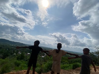 Three young boys stand on a dusty ridge overlooking a lush, green valley dotted with buildings and palm trees. The sky above is a brilliant blue, filled with dramatic, billowing clouds and a bright, shining sun. The boys, silhouetted against the sky, have their arms outstretched, seemingly embracing the vast landscape before them. The boy on the left wears a black t-shirt and shorts, the middle boy a light-colored tunic, and the boy on the right a dark grey t-shirt. The terrain they stand on appears to be a natural, unpaved embankment. The scene conveys a sense of freedom and wonder.