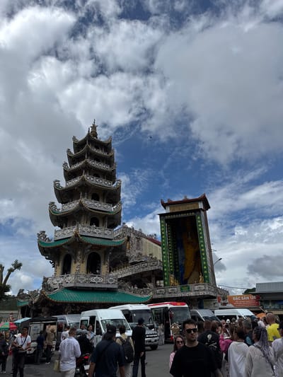 In Xuân Thành, Vietnam, a multi-story pagoda with green tiled roofs and ornate, detailed architecture stands tall under a cloudy sky. The pagoda features intricate carvings and multiple tiers, with a prominent tower to its left. In front of the pagoda, a busy street scene unfolds with several white vans and a small truck parked, suggesting a tourist or transportation hub. A crowd of people, diverse in age and attire, are gathered, some interacting with each other, others appearing to be tourists taking in the sights or waiting for transport. Some individuals are walking, while others stand near the vehicles. The overall atmosphere is one of a lively, public space with a significant cultural or religious landmark as the backdrop. A sign with Vietnamese text and the Coca-Cola logo is visible on a building to the right.