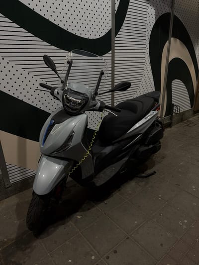 A light gray scooter is parked on a sidewalk in front of a wall with a black and white striped and dotted pattern. The scooter has a black seat, a clear windshield, and a yellow coiled cable attached to its front. The sidewalk is paved with gray square tiles. The scene is illuminated by artificial light, casting shadows on the ground. The location is identified as Vracar Urban Municipality, Serbia. No people are present in the image.