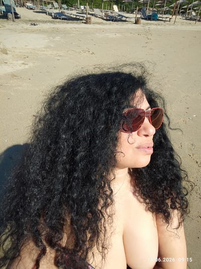 The image shows a woman with long, curly black hair and sunglasses on a sandy beach. She is wearing a bikini top and looking to the right. In the background, there are rows of beach chairs and umbrellas, suggesting a resort or public beach area. The timestamp in the lower right corner indicates the photo was taken on June 14, 2026, at 09:15. The location is identified as Rezovo, Bulgaria. The sand is light brown and appears dry. The lighting suggests it is daytime, possibly morning, with bright sunlight casting shadows.