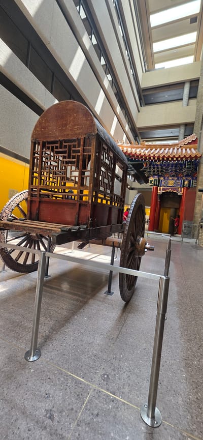 The image shows an ornate, wooden cart with decorative latticework on its sides and a curved wooden roof. It is supported by two large wooden wheels with spoked rims. The cart is positioned in what appears to be a museum or exhibition hall. In the background, there is a colorful, traditional-style structure with intricate carvings and vibrant colors, possibly representing an architectural element from East Asia. The floor is a speckled terrazzo with brass inlays. Sunlight streams in from above, casting shadows and highlighting the textures of the objects. Two metal support poles with circular bases are visible, holding up a horizontal metal bar that runs in front of the cart, suggesting it is part of an exhibit. The overall scene conveys a sense of historical display and cultural exhibition. The lighting suggests it is daytime. There is no visible text in the image.