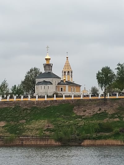 The image depicts a view of the city of Gorodnya in Russia. The main focus is a complex of religious buildings on a hill, likely a church or monastery, consisting of two prominent structures: one with a darker green roof and a golden dome topped with a cross, and another structure to its right with a bell tower. Both buildings appear to be of traditional Russian Orthodox architectural design.
The buildings sit atop a grassy hill with an artificial-looking, brown retaining wall at the bottom. A fence-like structure with white posts and a possible yellow railing runs along the top of the hill in front of the buildings.
The foreground shows a body of water, possibly a river or lake, with some vegetation along the shoreline.
The sky is overcast, suggesting a cloudy day. Trees are visible to the sides of the buildings, framing the scene.
