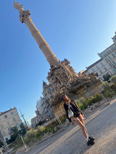A young woman with purple streaks in her hair, wearing a black hooded jacket, a pink skirt, fishnet tights, and white socks with black sneakers, stands on a brick-paved plaza. She is holding a metallic silver purse and looking at the camera. Behind her, a large, ornate stone column monument rises high into a clear blue sky. The monument features intricate sculptures of figures and animals at its base. In the background, several multi-story buildings with light-colored facades and green shutters line the street. The scene appears to be in a public square in Marseille, France, likely the Place de la Concorde or a similar grand plaza, given the architecture and the monument's style. The lighting suggests it is late afternoon or early morning.