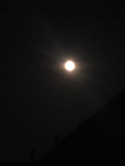 A full moon shines brightly in a dark sky over Khojniki, Belarus. The moon is positioned slightly left of center in the frame, casting a soft glow that illuminates thin wires stretched across the sky. In the lower right portion of the image, the dark silhouette of a building or structure is visible, its diagonal edge cutting across the frame. The overall scene is one of nocturnal stillness.