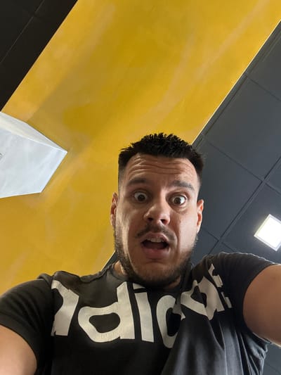 A man with dark hair and a beard, wearing a black t-shirt with a white logo, looks directly at the camera with wide eyes and an open mouth. He appears to be indoors, as indicated by the ceiling which is divided into sections of yellow and grey tiles, with a white light fixture visible. The man is positioned in the foreground, with his face taking up a significant portion of the frame. The perspective suggests the photo was taken from a low angle, looking upwards. The yellow section of the ceiling slopes downwards towards the viewer, and a white rectangular structure, possibly a vent or part of the architecture, is visible on the left side of the frame. The image does not contain any specific indicators of its location in Kyustendil, Bulgaria.