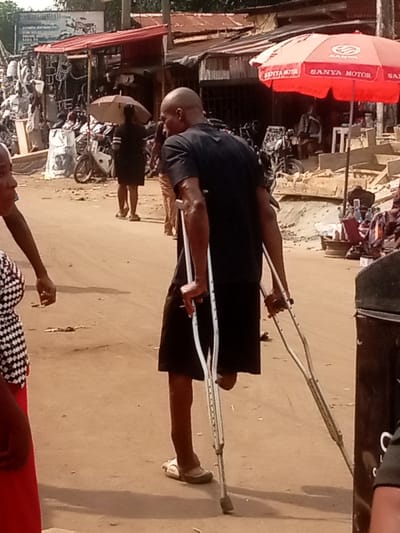The image displays a daytime street scene in a commercial area of Nnewi, Nigeria. The central figure, an adult male, is depicted from the rear-right as he walks towards the left side of the frame, utilizing two silver crutches. He is bald, wears a black short-sleeved shirt and black shorts. His right leg is amputated below the knee, and he wears a light-colored sandal on his left foot.
In the immediate foreground, on the far left, a person is partially visible, wearing a white and black checkered top and a red lower garment, facing left. In the mid-ground, a person in a dark dress holds a light-colored umbrella while walking away from the camera. The ground is an unpaved, light brown dirt surface.
To the left of the street, a series of market stalls and shops with corrugated metal roofs, some exhibiting rust, line the thoroughfare. These structures display a variety of goods, including what appear to be motorcycle parts and accessories. Several motorcycles are parked or positioned near these stalls. A sign with blue background and white lettering is visible above one shop. On the right side of the street, a large red umbrella branded with "SANYA MOTOR" provides shade over a vendor's area, where assorted items are visible on the ground or on low stands. A dark, rectangular object is partially visible in the extreme foreground on the right. The scene is brightly lit by natural light.