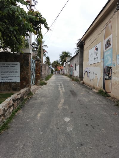 An eye-level, medium shot captures a narrow, asphalt-paved alleyway in Phan Thiet, Vietnam. The overcast sky suggests a cloudy day. The alley is flanked by buildings on both sides, with lush green trees and palm trees visible in the background, hinting at a tropical environment.
On the left side of the alley, a stone wall features a white sign with black text that reads, "WHY ARE YOU LITTERING? TAI SAO BAN XA RAC?" followed by several English and Vietnamese statements in smaller font, such as "I AM A JERK - TOI LA NGUOI NGO NGAN" and "I DON'T CARE ABOUT NATURAL AREAS - TOI CHANG QUAN TAM DEN MOI TRUONG." A blue metal gate is set into the wall.
The building on the right side of the alley is a light-colored structure adorned with various posters and graffiti. One prominent poster depicts a surfboard with the word "SURFPOINT" printed below it in blue lettering. Another poster features a circular logo with text. The wall itself has patches of peeling paint and what appears to be spray-painted artwork.
The asphalt road shows tire marks and scattered debris, including small dark stains and a few reddish-brown spots. The alley recedes into the distance, where more buildings and vegetation are visible. The scene appears to be a quiet, somewhat neglected urban area, possibly a residential or commercial side street.