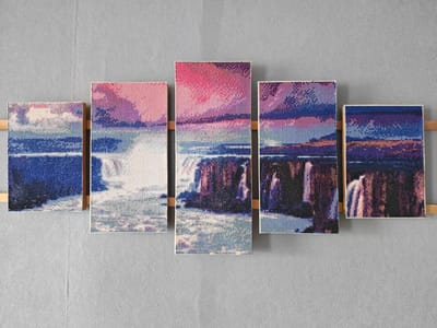 A set of five diamond paintings of a waterfall at dusk are mounted on a grey textured wall. The paintings are arranged horizontally and vary in size, with the two largest in the center and two smaller ones on either side, and one more on the far right. The paintings depict a cascading waterfall with water rendered in blues, whites, and purples. The sky above is a blend of pinks, purples, and blues, suggesting a sunset or sunrise. The scene is abstract, composed of small, faceted beads that create a pixelated effect. The paintings are framed with a light-colored wood and are attached to the wall with visible wooden supports. The overall impression is of a modern, decorative art installation. The location of this installation in Ashmyany, Belarus, is not specified in the image or its context.