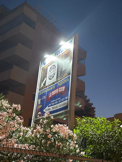 This is a nighttime photograph taken in Calp, Spain, featuring a brightly lit sign for "La mamma R0598," a restaurant and pizzeria. The sign is mounted on the side of a building, possibly a commercial establishment or apartment complex, which is partially visible in the background. The building has a modern architectural style with several floors.
The sign itself is predominantly blue and white and lists various food items like "Fritues," "Carnes," "Pescados," "Poke bowls," "Noodles," "Tacos," "Pastas," and "Pizzas." It also includes contact information: "Comida para llevar 647 063 400 - 617 921 799" and "150 Mts. Menús Variados." A small graphic of a woman or character is also present on the sign.
In the foreground, a dense green tree with vibrant pink flowers is visible, obscuring parts of the lower section of the sign and the building. The sky above is dark blue, indicating nighttime, and a faint beam of light emanates from the upper left corner of the image, possibly from a streetlamp or another light source. The overall impression is that of a lively evening scene in a Spanish town, with the restaurant sign being a focal point.