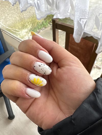 The image displays a left human hand, belonging to an adult with light skin tone, showcasing a fresh manicure. The scene is set indoors in the city of Dushevo, Bulgaria. The fingernails are almond-shaped and feature distinct designs: the thumb and pinky nails are solid white; the index finger nail has a yellow French tip; the middle finger nail is white with black speckles; and the ring finger nail is white with a yellow and white daisy-like floral pattern. The hand's wrist is covered by a dark, likely black, fabric sleeve. The background is out of focus, revealing a window with white patterned sheer curtains in the upper portion, through which blurred green foliage is visible outdoors. A partial view of a wooden chair with a brown frame is present to the right, and a dark blue object, possibly furniture, is visible in the lower left background. Natural light illuminates the subject from the window.
