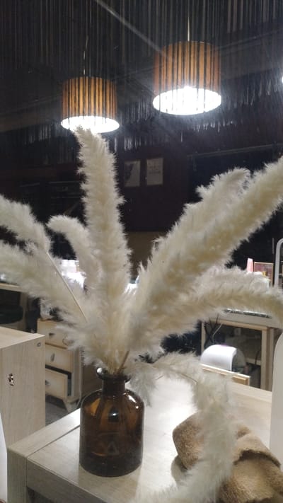 In Perea, Greece, an indoor scene depicts a cluster of dried, fluffy pampas grass stalks arranged in a dark amber glass vase. The vase rests on a light-colored wooden table. In the background, two pendant lights with slatted wooden shades hang from a dark ceiling. The environment appears to be a room with some furniture, including a light-colored cabinet with drawers to the left and what looks like a desk or workbench with equipment in the mid-ground to the right. The lighting is dim, with the pendant lights providing the primary illumination. There are no people visible in the image.