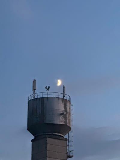 This is a vertical, low-angle, outdoor, daytime photograph taken in the city of Cherven, Belarus. The image features a water tower with a concrete base and a metal tank on top. A metal railing encircles the top of the tank. A rooster-shaped weather vane is positioned on the railing, and two rectangular antenna panels are mounted on the tower. A ladder runs up the side of the tank. A crescent moon is visible in the pale blue sky, which is partially obscured by clouds. The scene appears to be set during twilight.