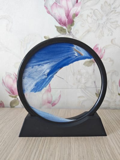 This media file features a black, circular, kinetic sand art frame placed on a light-colored wooden surface. The frame is mounted on a black, angled base. Inside the frame, a landscape of blue and white sand is depicted, with the sand having shifted to create undulating patterns and peaks. The background is a textured wallpaper adorned with a floral pattern of pink magnolias and subtle light gray swirling designs. The lighting appears to be from an overhead source, casting soft shadows. No people are visible in the image. The scene appears to be indoors, on a flat surface, with a decorative background. The image itself is a still shot, with no discernable activity taking place beyond the static arrangement of the sand art. There is no visible text in the image.