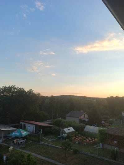 The image shows a rural scene in Dobev, Czechia, likely during the late afternoon or early evening, indicated by the soft, warm light and the colors in the sky. The foreground features a residential area with several structures and outdoor amenities. To the left, a green umbrella with a beer logo, "Pilsner Urquell", is set up over a patio table and chairs, suggesting an outdoor seating area. Adjacent to this is a small building with a red roof. Further into the scene, there are greenhouses, raised garden beds, a clothesline, and other smaller outbuildings. Behind the immediate residential area, a dense forest stretches across the landscape, with rolling hills visible in the distance. The sky is a clear blue with scattered white and pale orange clouds. No people are visible in the image.