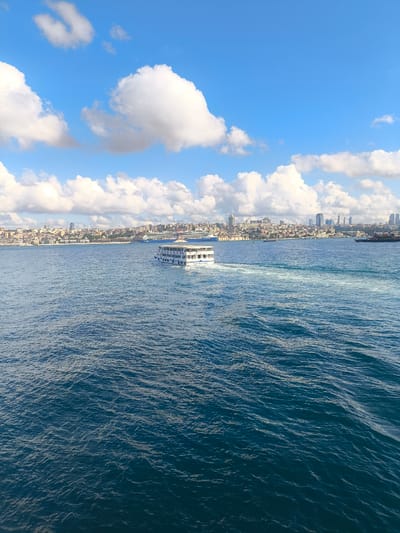 The image depicts a wide view of the Bosphorus strait in Üsküdar, Turkey, on a clear day with scattered clouds. The foreground is dominated by the deep blue, rippling water of the strait. A white passenger ferry with multiple decks is traveling from left to right across the mid-ground, leaving a white wake behind it. In the background, the cityscape of Istanbul stretches along the coastline, characterized by a dense collection of buildings, including residential areas, commercial structures, and skyscrapers. The sky is a vibrant blue, with large, fluffy white clouds adding texture and depth to the scene. The overall impression is one of a busy urban waterway against a backdrop of a bustling city.