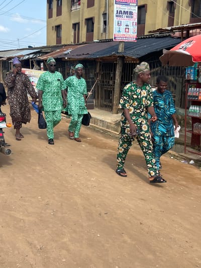 The scene depicts a street in Ijegun, Nigeria. Four men in traditional attire are present. Two are sitting in front of a shop, while the other two are walking along the road. There is a banner advertising an academy for JAMB/WAEC/NECO exams. The buildings are simple structures. A motorcycle is parked on the side of the road. The road is unpaved.