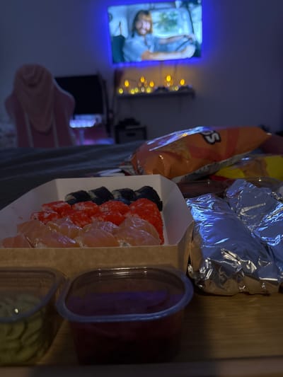A still life, likely indoors, shows a take-out sushi order, a bag of chips, and other snacks on a surface. The sushi is in a white cardboard container with a variety of rolls, including those topped with salmon, tobiko (flying fish roe), and seaweed. Sauces are in clear plastic containers beside the sushi. A partially obscured bag of Doritos chips is behind the sushi. Silver foil-wrapped food is partially visible. In the background, a television with a blue glow displays a video. Fairy lights are draped nearby. An object resembling a chair or clothing item is partially visible.

There is no information in the image to suggest the location is in the city of Polje, Montenegro.