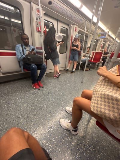 The image depicts the interior of a subway car in Nuremberg, Germany. Several people are visible, some seated and others standing. One woman in the foreground is wearing a black tank top and denim shorts, with a blue baseball cap on her head. Another woman, partially visible, is holding a checkered bag and wearing white sneakers. A man in a light blue denim shirt is seated and looking at his phone. A person in dark pants and red sneakers is also seated. The subway car has white walls, red seats, and a gray speckled floor. Overhead poles and handles are present for passengers to hold onto. The lighting suggests it is daytime. The scene captures a typical commute, with individuals engaged in their own activities. There is no visible text in the image.