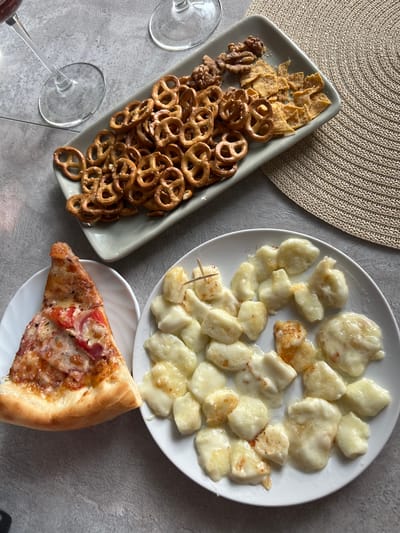 A high-angle, medium shot captures a spread of snacks and drinks on a textured gray surface, potentially a table, in Magnitogorsk, Russia. To the upper left, two wine glasses are visible, one partially filled with a dark liquid, suggesting wine. A rectangular light gray tray occupies the upper center of the frame, filled with a pile of mini pretzels, some shelled walnuts, and a scattering of crunchy corn chips. Below and to the left of the tray, a white plate holds a single slice of pizza, topped with melted cheese, tomato, and what appears to be ham. To the right of the pizza, a round white plate is covered with small, irregularly shaped pieces of what looks like fried or baked cheese, possibly halloumi or a similar variety, lightly seasoned with red pepper flakes. The overall scene suggests a casual gathering or a relaxed meal.