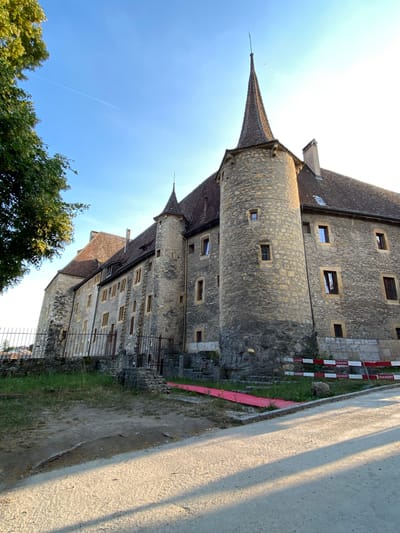 The image depicts a historic stone castle building with a conical roofed turret and a tall, pointed spire in the city of Milvignes, Switzerland. The castle, constructed from rough-hewn stone blocks and featuring numerous windows with dark frames, is situated on a grassy slope. A black metal fence runs along the front of the property, partially obscured by a tree with green leaves on the left side of the frame. Construction barriers, white with red lettering that reads "DUCKERT", are visible in front of the castle, and a red pathway or covering is laid on the ground leading towards the building. The sky is clear and blue, with a faint contrail from an aircraft visible towards the upper left. The ground in the foreground is a dirt path with shadows cast by the setting sun.