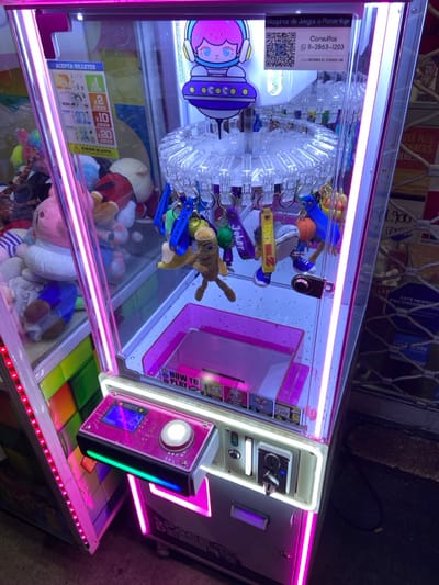 A "Space Debris" arcade prize machine, prominently lit by internal pink and white LED strips, is positioned in a commercial environment in San Antonio de Padua, Argentina. The machine's transparent acrylic casing reveals a central, clear plastic carousel dispensing multiple keychains as prizes, including depictions of "Joker," "Chucky," a banana, and an alien-like character. A graphic of a character in a spacesuit, marked "COCO," is mounted above the prize carousel. An instructional decal states "ACEPTA BILLETES" (Accepts Bills) and details game pricing (1, 2, 10, 20 games for 1000, 2000, 5000, 10000 currency units, respectively), alongside a QR code and contact number "11-2863-1203" for "Consultas" (Inquiries). Below the prize mechanism, a clear plastic retrieval chute is labeled "HOW TO PLAY." The machine's control panel includes a blue-lit display screen, a large, illuminated white push-button, and a coin/bill acceptor slot, all accented with rainbow-colored LED strips. To the left, another arcade machine containing plush toys is partially visible. The background on the right features a partially lowered metal security gate and obscured signage displaying "1900" and fragmented text, including "ESTE NEGOCIO PIDE DOCUMENT." No individuals are present.
